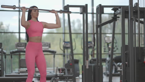 Hispanic-brunette-woman-in-a-pink-suit-pushes-a-barbell-over-her-head-to-the-top-while-training-her-shoulders-in-the-gym.-Standing-exercise-for-training-the-shoulders-and-arms.-Weight-training
